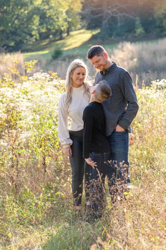 Family of three with blond mom, tall dad, and 10 year old son looking up at his parents in wild, grassy field with sunny backlight